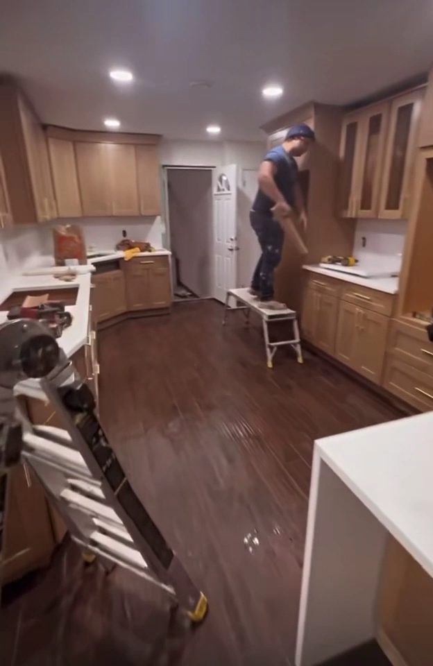 An installer on a step stool fitting wall cabinets in a kitchen with dark hardwood flooring already laid.