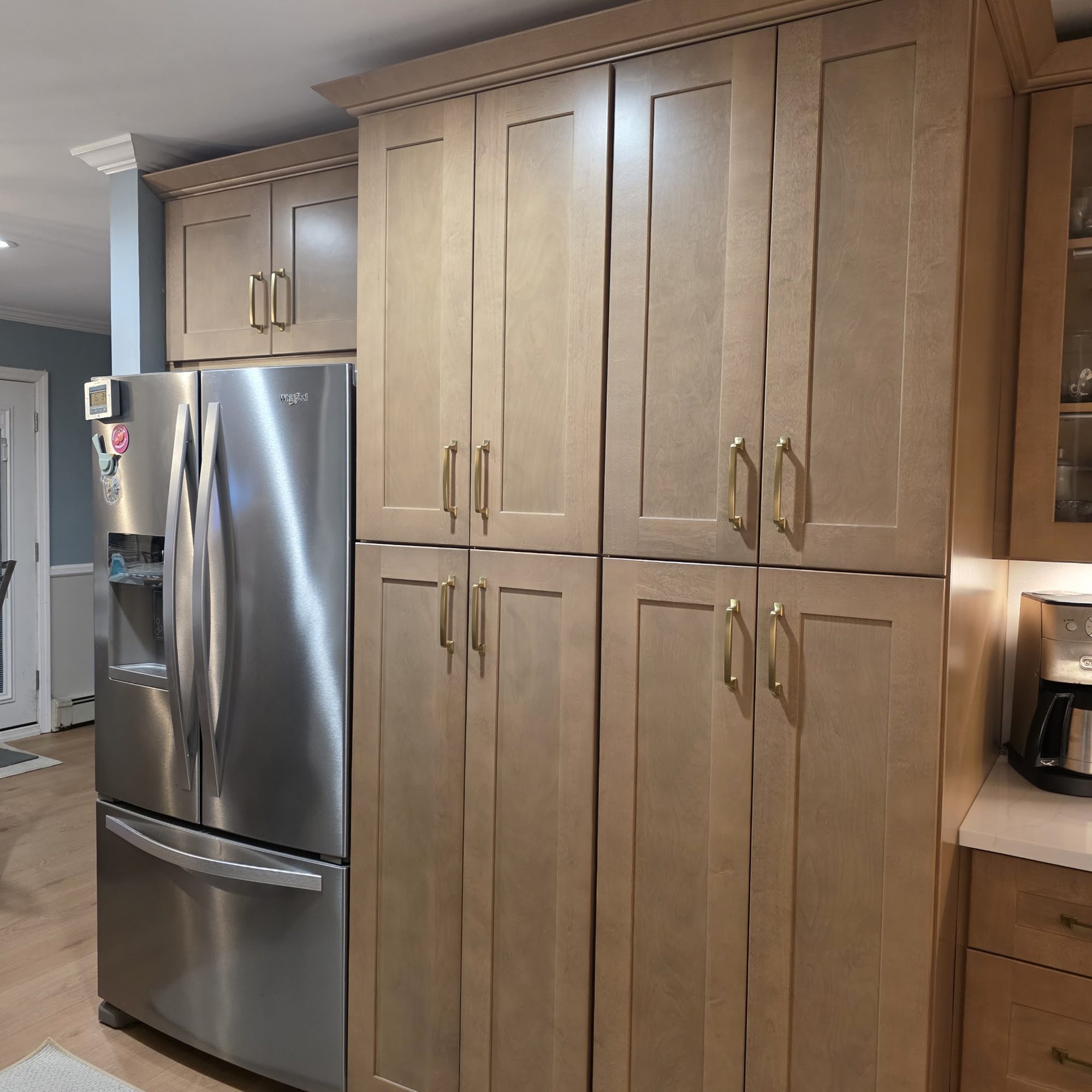 A full-height maple shaker pantry wall with brushed gold handles beside a stainless refrigerator.
