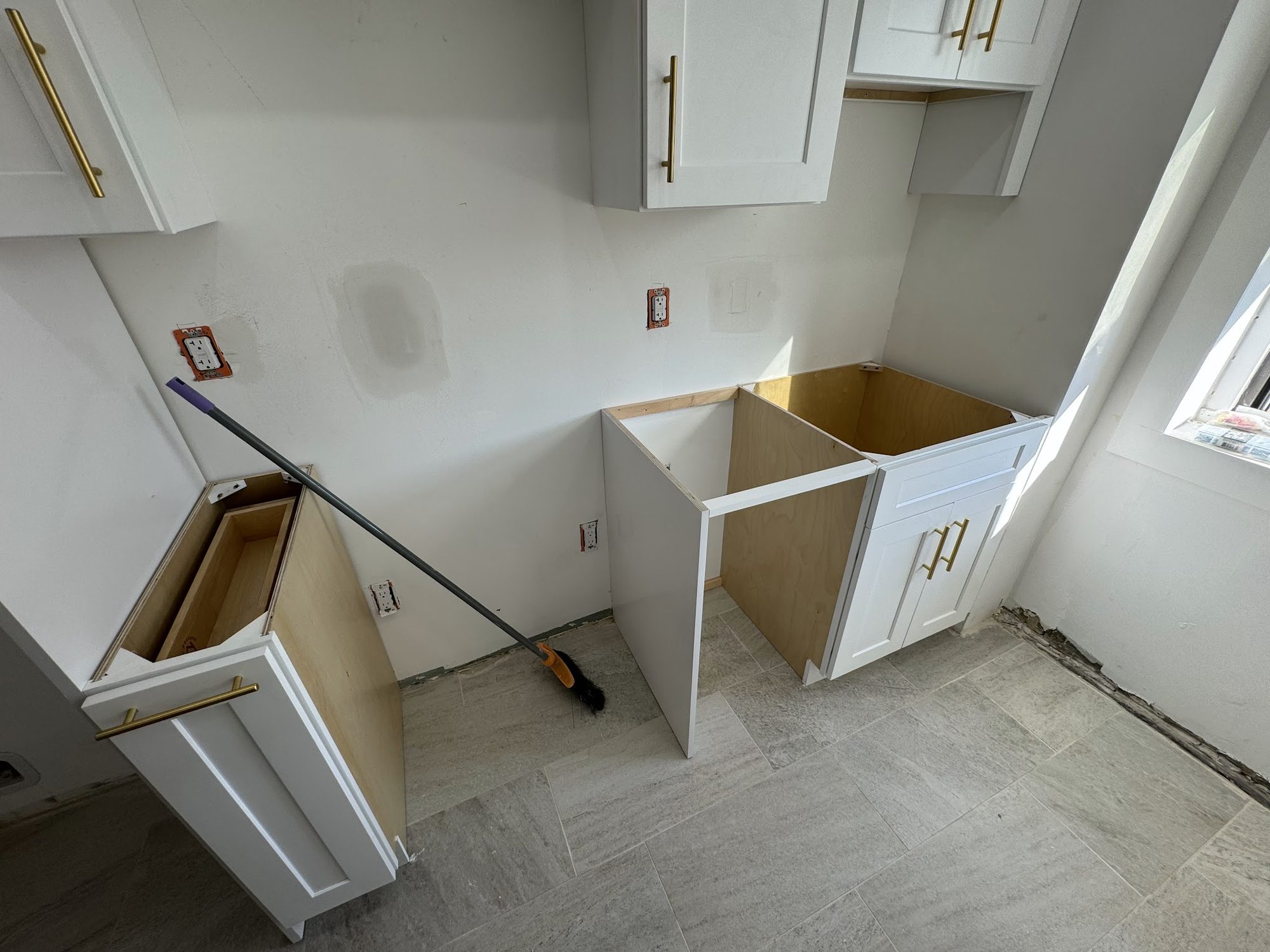 White shaker cabinets with brushed gold bar pulls being installed over grey tile flooring.